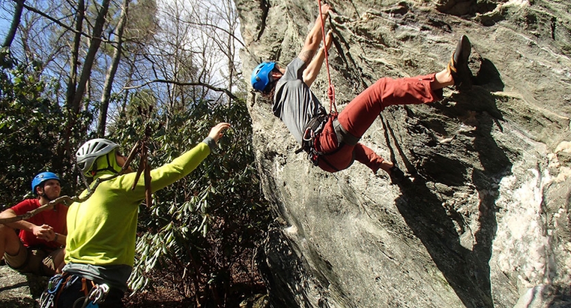 A person wearing safety gear is secured by ropes as they rock climb. Two others look on from the ground. 